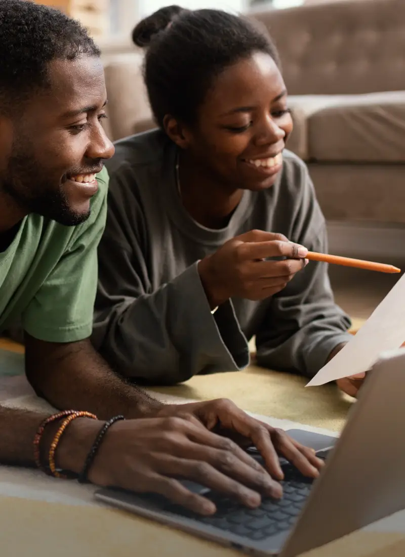 Two individuals conducting a collaborative valuation analysis at home, using a laptop and paper while lying on a rug in a cozy living room, discussing financial data or project planning.